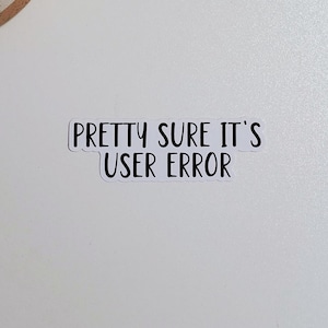 May include: A white sticker with the text "PRETTY SURE IT'S USER ERROR" in black, placed on a white surface. A small potted cactus is in the upper left corner.