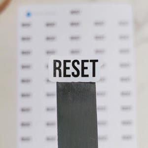 May include: A white sticker with the word "RESET" in black capital letters is placed on a metal tool. The sticker is in front of a sheet of small white stickers. The background is a white surface.