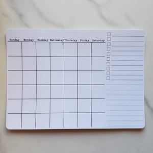 May include: A white rectangular planner with a grid for the days of the week and checkboxes. The days of the week are listed across the top, with space for notes and tasks on the right side. The planner is on a white marble surface.