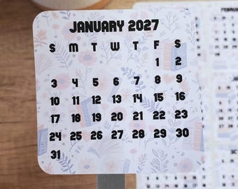 May include: A square sticker featuring a January 2027 calendar with a light background patterned with small blue and pink flowers and books. The text JANUARY 2027 is printed in bold black letters at the top, followed by the days of the week and dates.