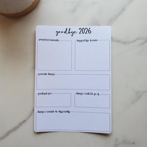 May include: A white planner with the text "goodbye 2026" in a handwritten style. The planner includes prompts for "proudest moments," "biggest life lessons," "favorite things," "grateful for," and "things I will let go of."