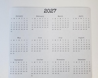 May include: A white rectangular 2027 calendar with rounded corners, featuring twelve monthly grids arranged in three rows. Each month displays the days of the week abbreviated as S, M, T, W, T, F, S, with black printed numbers for each date.