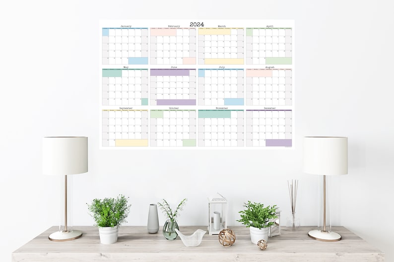 Op de afbeelding: Een witte muur met een 2024-kalender erop gedrukt. De kalender is verdeeld in 12 maanden, elk met een andere pastelkleur. De kalender hangt aan de muur boven een houten tafel met twee lampen, een vaas met bloemen en een decoratieve lantaarn.