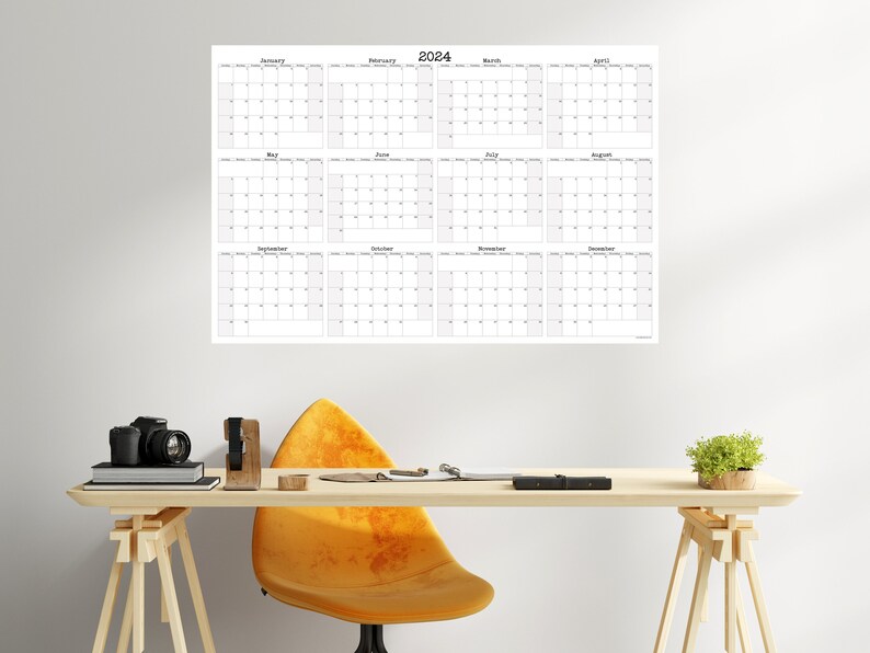 Op de afbeelding: Een zwart-witte 2024-kalenderposter met 12 maanden weergegeven in een rasterformaat. De poster hangt aan een witte muur boven een houten bureau met een gele stoel, een camera en een potplant.