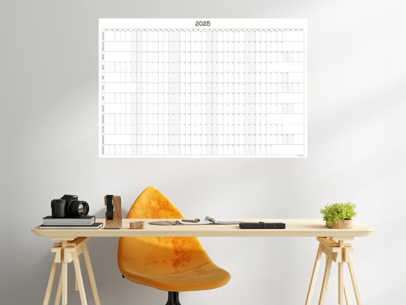 May include: A white wall calendar with the year 2025 printed at the top. The calendar is divided into months and days, with each day having a small square for writing in appointments or events. The calendar is hanging on a white wall.