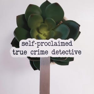 May include: A white sticker with black text that reads "self-proclaimed true crime detective" is attached to a metal plant stake. The stake is inserted into a green succulent plant.