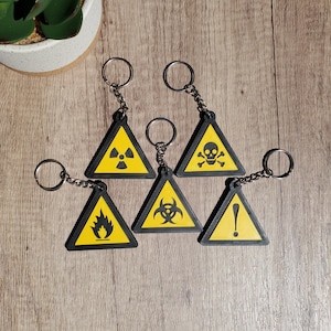 May include: Set of six black and yellow triangle-shaped keychains with different hazard symbols. The symbols include a skull and crossbones, a radiation symbol, a biohazard symbol, an exclamation point, and a flame.