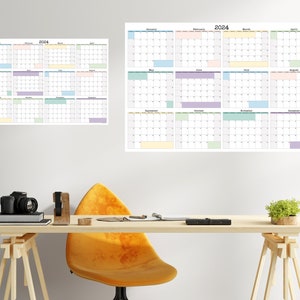 Op de afbeelding: Een bureau met een gele stoel, een camera en een plant. Aan de muur hangen twee kalenders, een voor 2024 en een voor 2023. De kalenders zijn in een rasterformaat met elke maand weergegeven in een apart vak. De maanden zijn kleurgecodeerd met verschillende pastelkleuren.