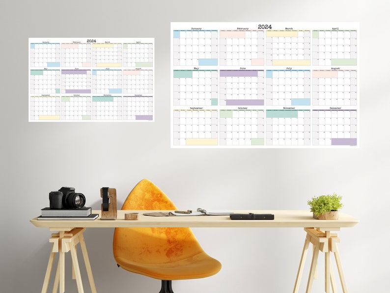 Op de afbeelding: Een bureau met een gele stoel, een camera en een plant. Aan de muur hangen twee kalenders, een voor 2024 en een voor 2023. De kalenders zijn in een rasterformaat met elke maand weergegeven in een apart vak. De maanden zijn kleurgecodeerd met verschillende pastelkleuren.