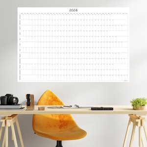 May include: A white wall calendar with the year 2024 printed at the top. The calendar is hanging on a white wall above a wooden desk with a yellow chair, a camera, and a potted plant.