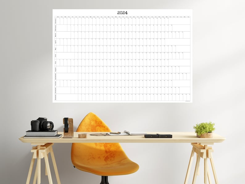 May include: A white wall calendar with the year 2024 printed at the top. The calendar is hanging on a white wall above a wooden desk with a yellow chair, a camera, and a potted plant.