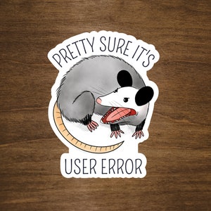 May include: A white sticker with a cartoon possum with a black nose and black eyes. The possum is snarling and has a pink tongue sticking out. The text "Pretty sure it's user error" is written in black around the possum.