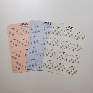 May include: Three calendars, one for each year 2024, 2025, and 2026. The calendars are printed on different colored paper: peach, light blue, and cream. Each calendar has a grid layout with the days of the week labeled at the top and the numbers for each day of the month listed below.