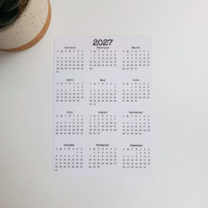 May include: A white rectangular 2027 calendar card featuring twelve months arranged in a grid. Each month displays the days of the week and corresponding dates in a black typewriter-style font. The calendar is printed on a smooth, flat surface.