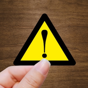 May include: A yellow triangle warning sign with a black exclamation point in the center. The sign is being held by a hand.