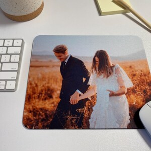 May include: A couple in wedding attire walks through a field of tall grass. The groom is wearing a dark suit and the bride is wearing a white dress. The background is a hazy sunset.