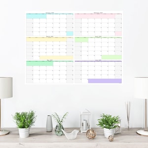 May include: A wall calendar with six months displayed, January through June. Each month is color-coded with a different pastel color. The calendar is hanging on a white wall above a table with two lamps, plants, and decorative items.