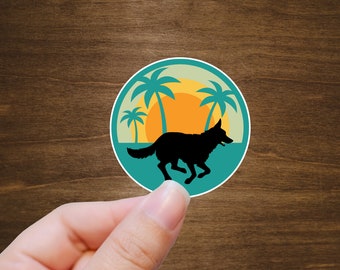 May include: A round sticker with a teal background and a silhouette of a dog running on a beach with palm trees and a sunset in the background.
