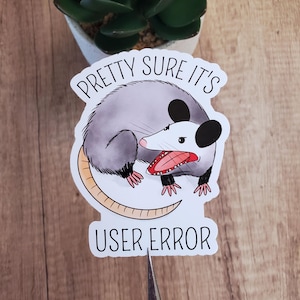 May include: A die-cut sticker featuring a gray and white opossum with a pink nose and ears, showing its teeth. The text above reads PRETTY SURE IT'S and the text below reads USER ERROR in a black sans-serif font against a white background.