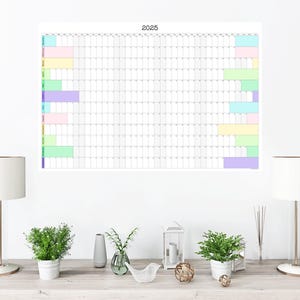 May include: A white wall with a 2025 calendar poster hanging on it. The calendar has a grid layout with colored bars indicating different events. Two white lamps with white lampshades are on a wooden table in front of the wall. There are also two potted plants and a decorative bird figurine on the table.