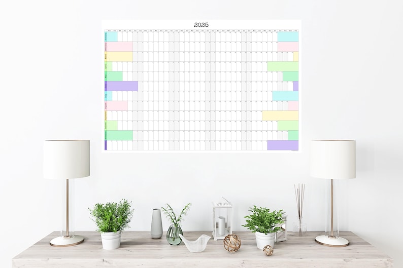 May include: A white wall with a 2025 calendar poster hanging on it. The calendar has a grid layout with colored bars indicating different events. Two white lamps with white lampshades are on a wooden table in front of the wall. There are also two potted plants and a decorative bird figurine on the table.