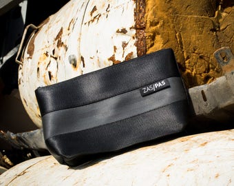 Può includere: Una pochette nera con cerniera, con una striscia grigia e il testo "ZAS/PAS" sul davanti. La pochette è appoggiata su una superficie metallica arrugginita.