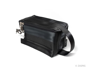 May include: Black zippered travel bag made from recycled seatbelt material. The bag has a black strap and a small black tag with the word "ZASPAS" printed on it.