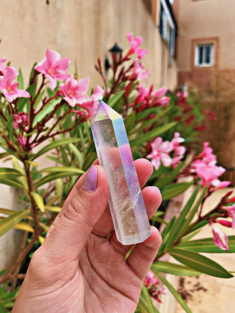 May include: A clear crystal point with iridescent rainbow colors. The crystal is held in a hand against a background of pink flowers.
