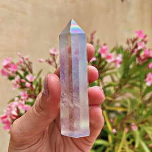 May include: A clear iridescent crystal point with a rainbow sheen. The crystal is held in a hand against a background of pink flowers and green foliage.