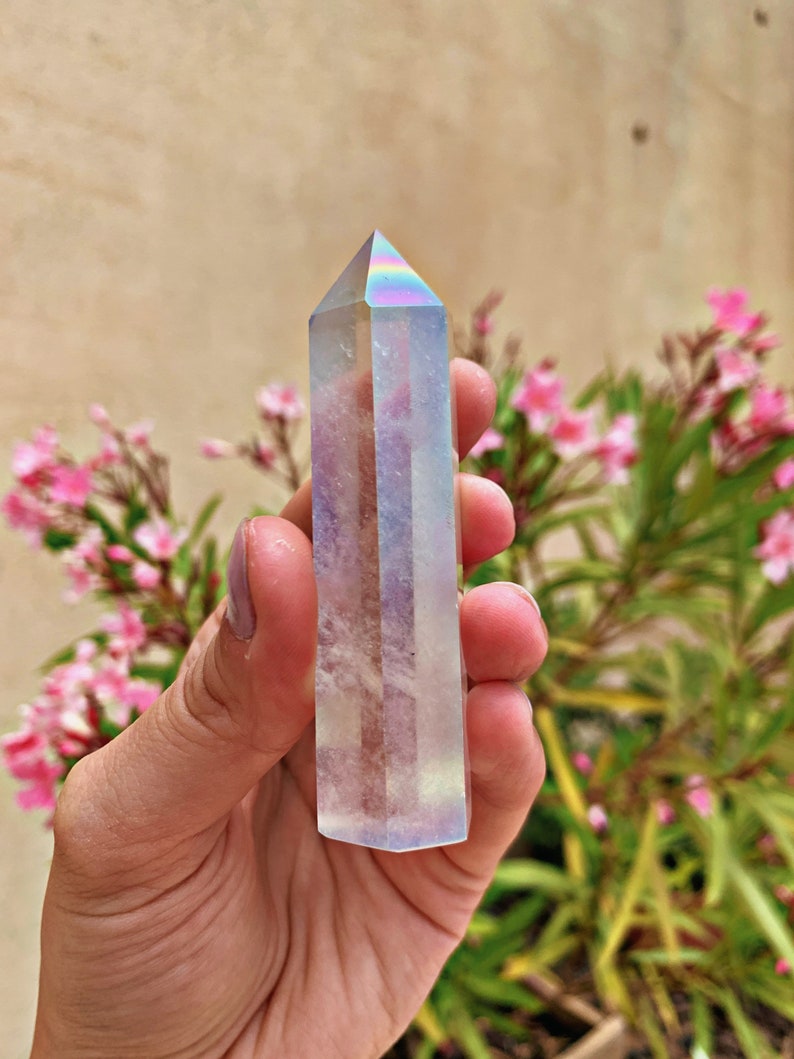 May include: A clear iridescent crystal point with a rainbow sheen. The crystal is held in a hand against a background of pink flowers and green foliage.