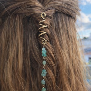 Könnte beinhalten: Goldfarbene Haarspirale mit einer Kette aus kleinen, hellblau-grünen Edelsteinen. Die Spirale ist in einen Zopf aus hellbraunem Haar eingesetzt und bildet ein dekoratives Haaraccessoire.