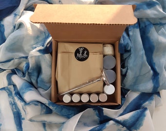 Pode incluir: Uma caixa de cartão castanho com a tampa aberta, revelando um conjunto de seis pequenos recipientes redondos, uma ferramenta de metal e um saco de papel castanho com as palavras "Obrigado" impressas nele. A caixa está sentada sobre um tecido estampado azul e branco.
