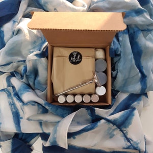 May include: A brown cardboard box with a lid open, revealing a set of six small round containers, a metal tool, and a brown paper bag with the words "Thank You" printed on it. The box is sitting on a blue and white patterned fabric.