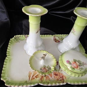 Könnte beinhalten: Ein Vintage-Porzellan-Waschtisch-Set mit einem rechteckigen Tablett, zwei Kerzenhaltern, einer kleinen Schale und einer Schmuckdose mit Deckel. Das Set ist weiß mit grünen Verzierungen und Rosenblüten-Akzenten. Die Kerzenhalter sind etwa 15 cm hoch.