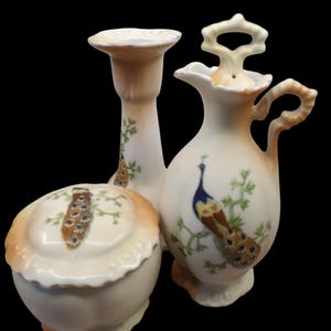 May include: A vintage porcelain bathroom set featuring a peacock design. The set includes a lidded jar, a pitcher, and a candle holder.