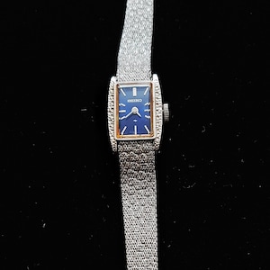 May include: A vintage Seiko wristwatch with a rectangular blue face and silver-tone metal strap. The watch face has silver hands and markers. The strap is a fine mesh design. The watch is a classic style.