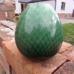 May include: A green, egg-shaped ceramic vase with a diamond pattern. The vase is glossy and rests on a red brick wall. The background includes a house and green grass.