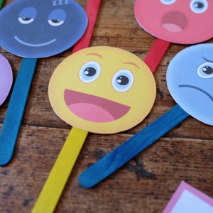 May include: A set of six colorful paper face masks with different expressions, including sleepy, happy, sad, and surprised. Each mask is attached to a wooden stick.