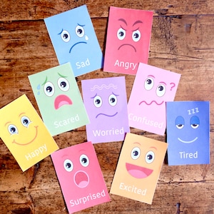 May include: A selection of brightly coloured cards, each illustrating a different emotion with a matching facial expression and word label. Emotions include happy, sad, angry, scared, worried, confused, excited, surprised, and tired. The cards are on a wooden surface.
