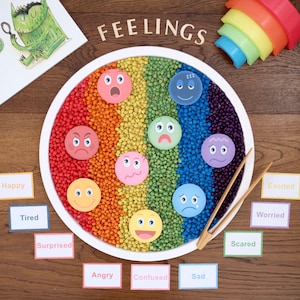 May include: A white tray filled with colorful beads arranged in a rainbow pattern.  The tray has eight different emotion faces on it.  The words "FEELINGS" are written in wooden letters above the tray.  There are 10 cards with different emotions written on them, such as "Happy", "Tired", "Surprised", "Angry", "Confused", "Sad", "Excited", "Worried", "Scared", and "Angry".