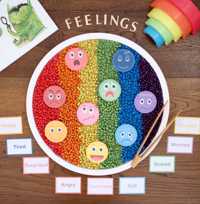 May include: A white tray filled with colorful beads arranged in a rainbow pattern.  The tray has eight different emotion faces on it.  The words "FEELINGS" are written in wooden letters above the tray.  There are 10 cards with different emotions written on them, such as "Happy", "Tired", "Surprised", "Angry", "Confused", "Sad", "Excited", "Worried", "Scared", and "Angry".