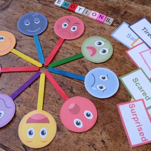 May include: A set of eight colorful paper face masks with different facial expressions, including happy, sad, angry, surprised, scared, sleepy, and confused. The masks are attached to wooden sticks and arranged in a circle on a wooden surface. The word "EMOTIONS" is spelled out in colorful letter tiles.