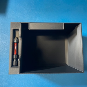 Puede incluir: Una caja de almacenamiento de plástico negro con un pequeño compartimento para una punta de destornillador. La caja está vacía y la punta del destornillador es visible.