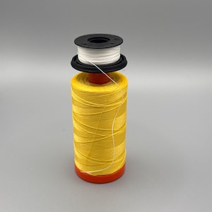 Puede incluir: Una bobina de hilo amarilla con una canilla blanca en la parte superior. La bobina está sobre una superficie gris.