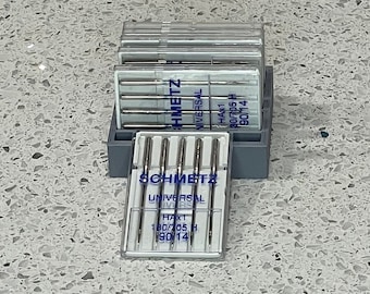 May include: A clear plastic container with a gray lid holds a set of sewing machine needles. The needles are labeled "SCHMETZ UNIVERSAL HAX1 180/705 H 90/14".