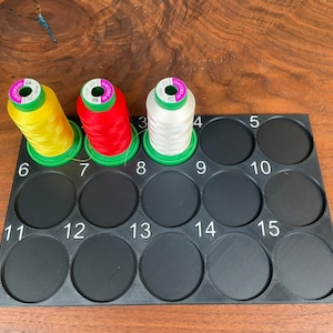 May include: A black plastic thread organizer with 15 numbered compartments. Three spools of thread are in the compartments, one yellow, one red, and one white.