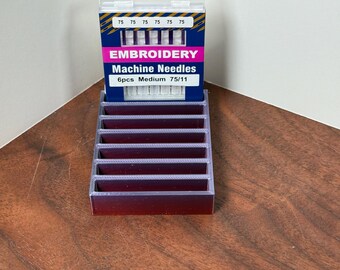 May include: A package of embroidery machine needles, labeled "Embroidery Machine Needles 6pcs Medium 75/11". The package is blue and white with a clear plastic hanger. The needles are displayed in a clear plastic organizer.