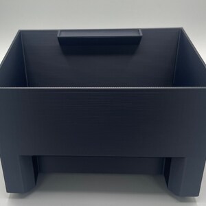 Puede incluir: Una caja de almacenamiento de plástico azul oscuro con un pequeño compartimento en la parte superior. La caja tiene un recorte en la parte delantera.