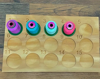 May include: A wooden thread organiser with 15 numbered compartments. Four spools of thread are in the compartments, one pink, one blue, one turquoise, and one light blue.