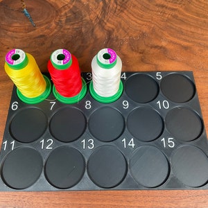 May include: A black plastic thread organiser with 15 numbered compartments. Three spools of thread are in the compartments, one yellow, one red, and one white.
